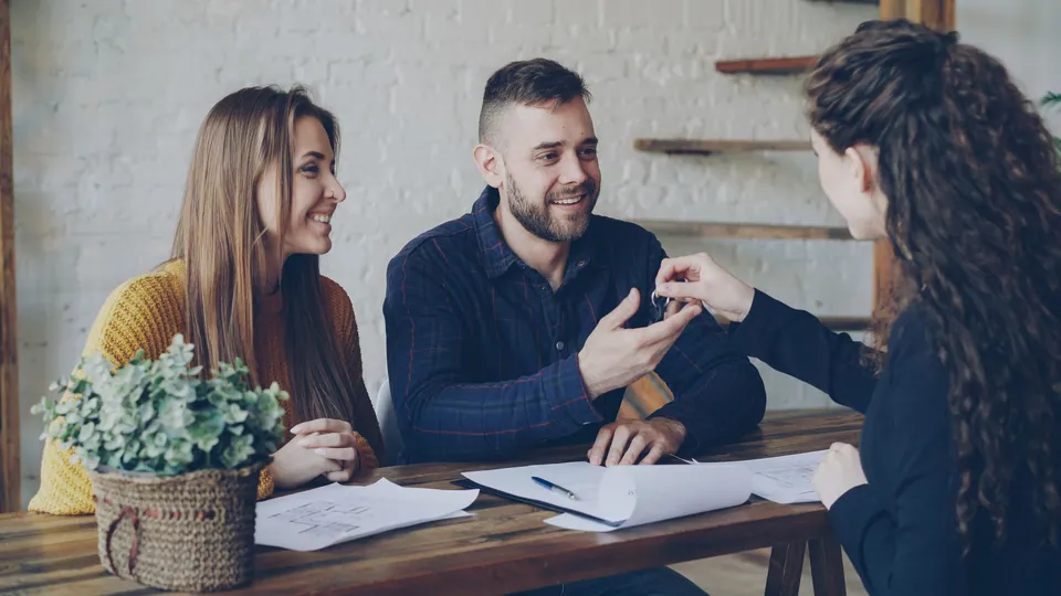A smiling couple receiving a set of keys from a property agent across a table with floor plans